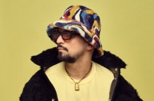Person wearing a colorful bucket hat, sunglasses, gold necklace, and a black fuzzy jacket against a yellow background.