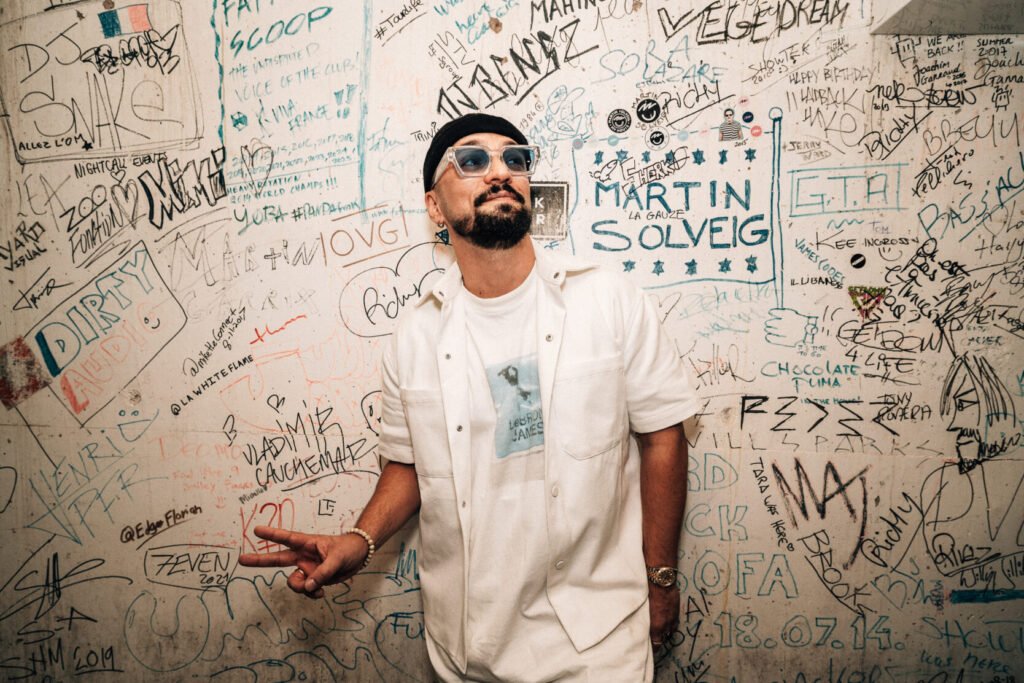 Dombresky in a white outfit and sunglasses poses with a peace sign in front of a graffiti-covered wall.