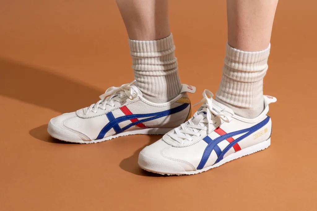 White athletic shoes with blue and red stripes, worn with cream-colored socks on a brown floor.