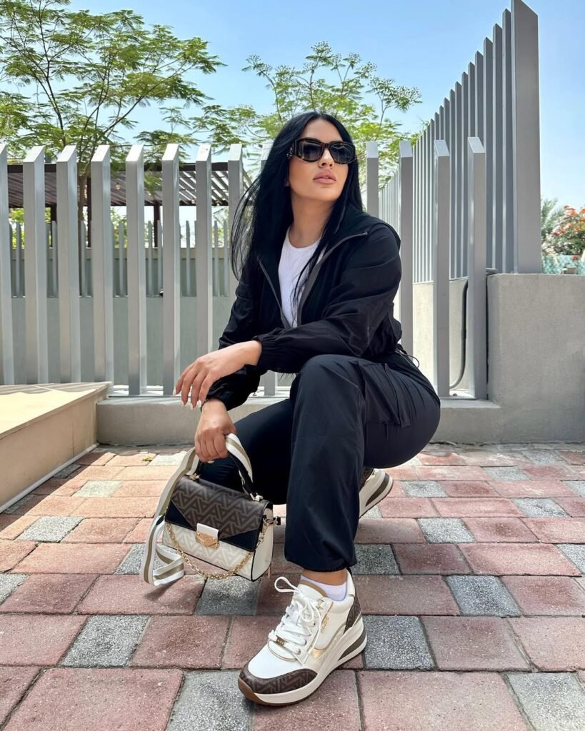 A woman with long black hair, wearing sunglasses and a black outfit, crouches on a paved walkway with a patterned bag. Global Sneaker Testing