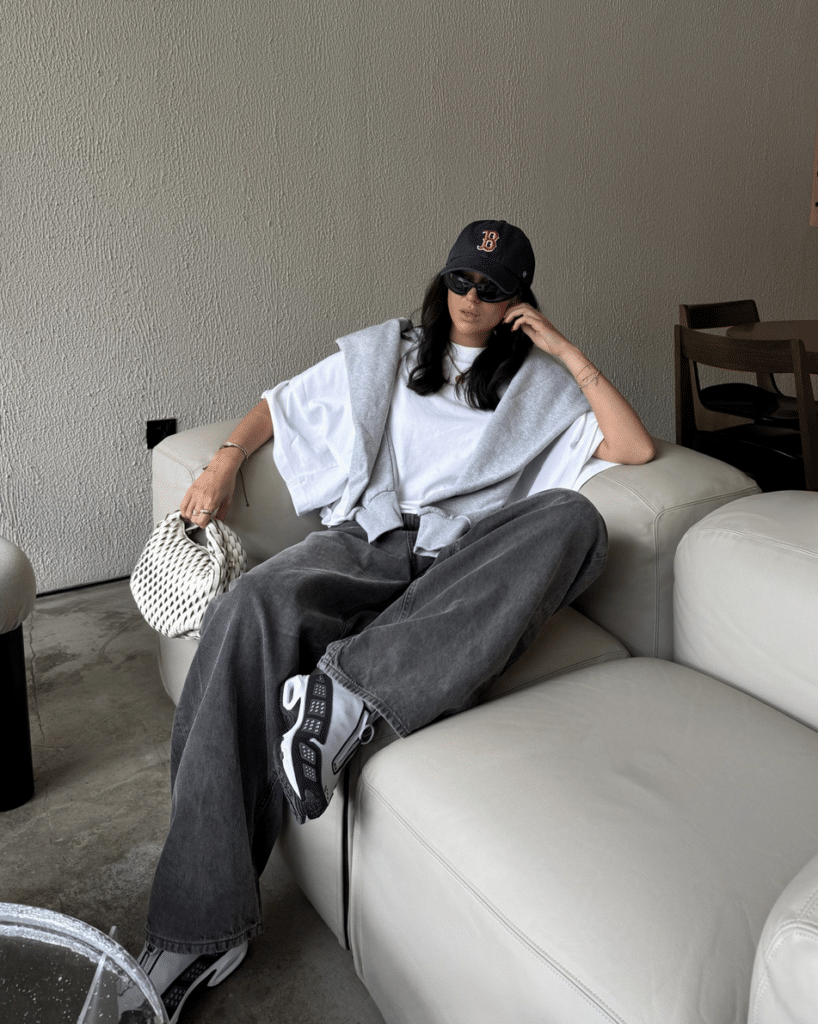 A person in a white t-shirt, gray sweater, dark pants, and a cap, relaxes on a cream couch with sunglasses and a woven bag. Global Sneaker Testing.