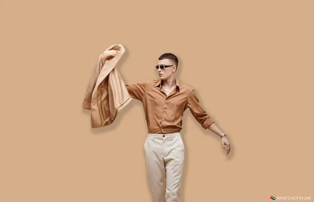 A man wearing sunglasses and a brown shirt, holding a beige jacket against a beige background.