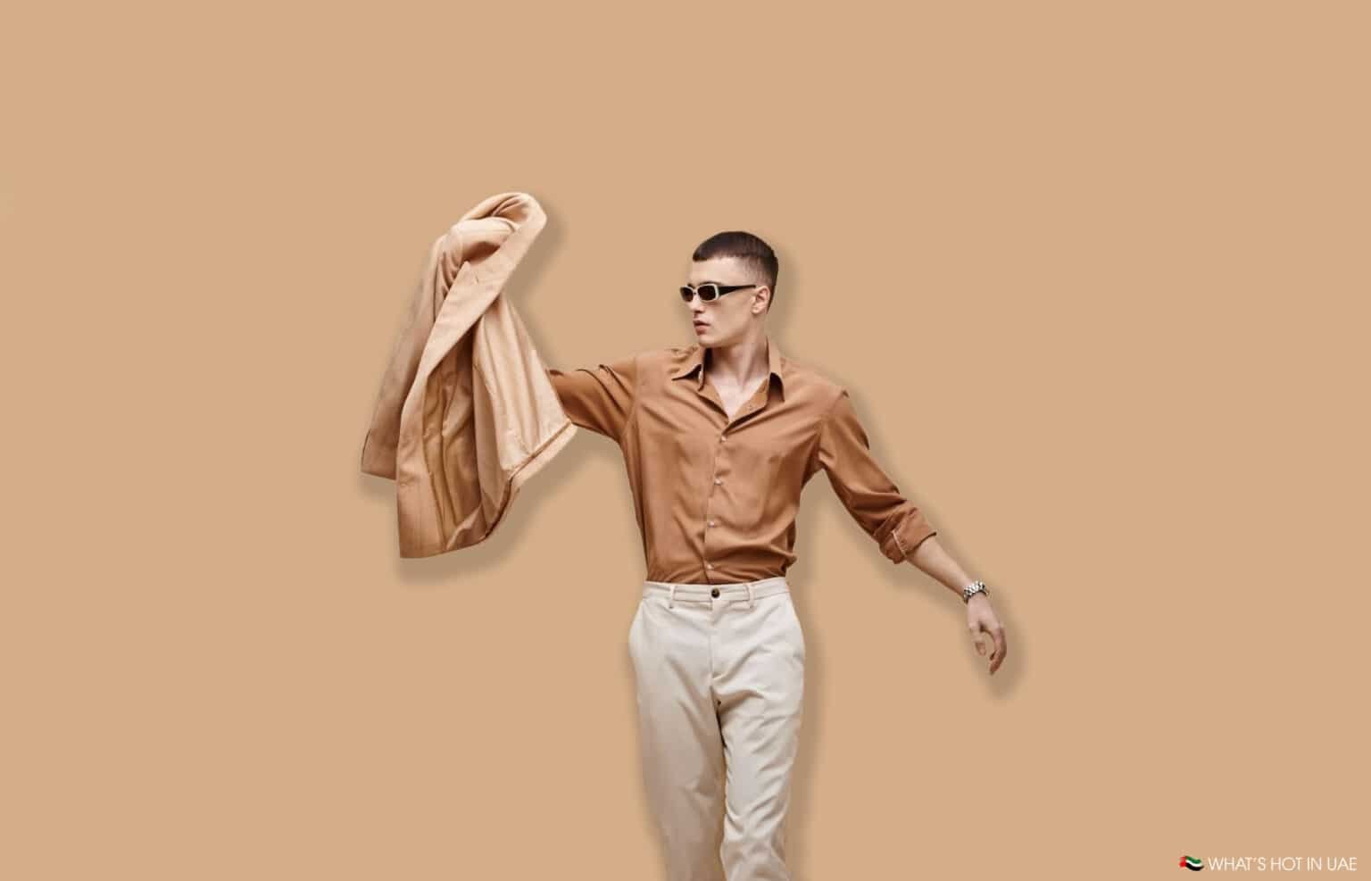 A man wearing sunglasses and a brown shirt, holding a beige jacket against a beige background.