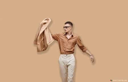 A man wearing sunglasses and a brown shirt, holding a beige jacket against a beige background.