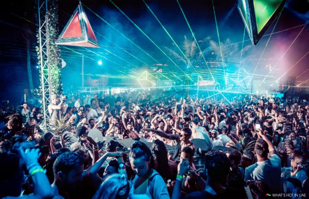 A vibrant crowd dances at a nighttime outdoor party, illuminated by colorful lasers and geometric light displays.