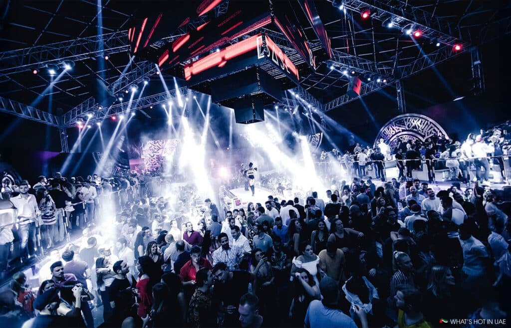 A crowded nightclub scene with colorful lights illuminating a large dance floor filled with people.