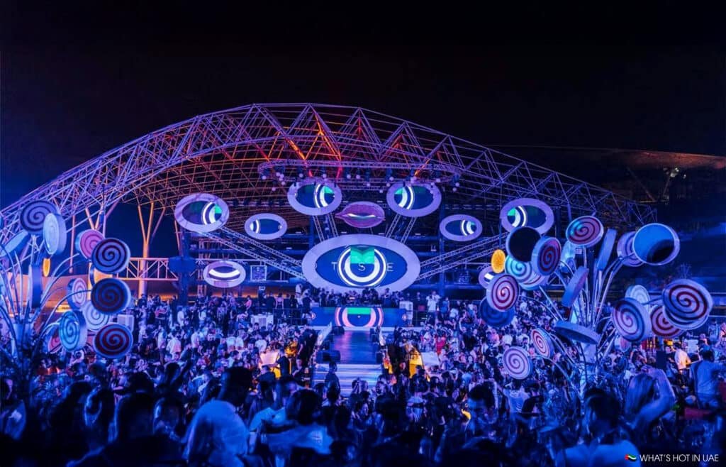 A vibrant outdoor concert with a large stage featuring circular screens, surrounded by a lively Dubai Nightlife crowd at night.