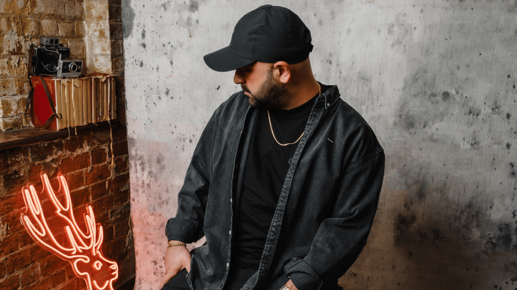 A man in a black outfit and cap sits beside a glowing deer-shaped neon light, against a brick and concrete wall backdrop.