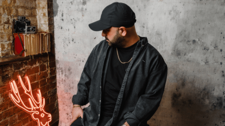 A man in a black outfit and cap sits beside a glowing deer-shaped neon light, against a brick and concrete wall backdrop.