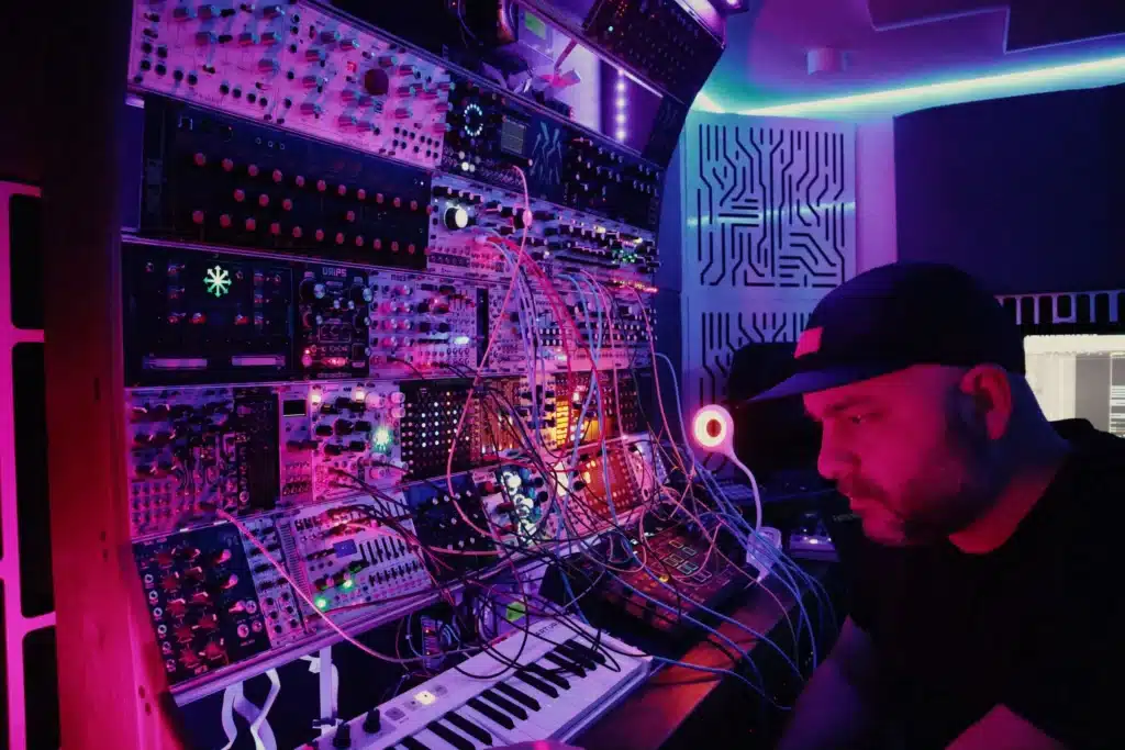 A person wearing a cap is sitting beside a large array of brightly lit synthesizer modules and a keyboard in a dimly lit room.