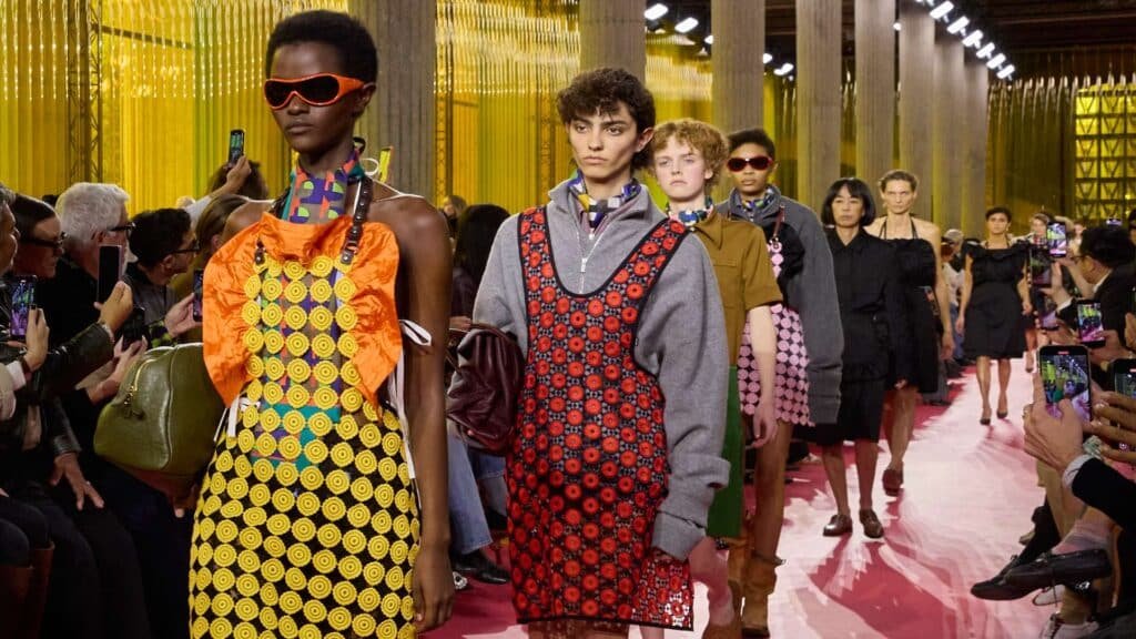 Models showcase colorful outfits on a runway during a fashion show, surrounded by an audience taking photos.