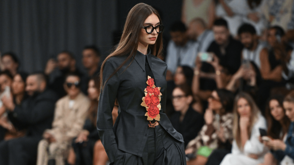 A model in a dark outfit adorned with red roses walks on a runway; the audience is blurred in the background.
