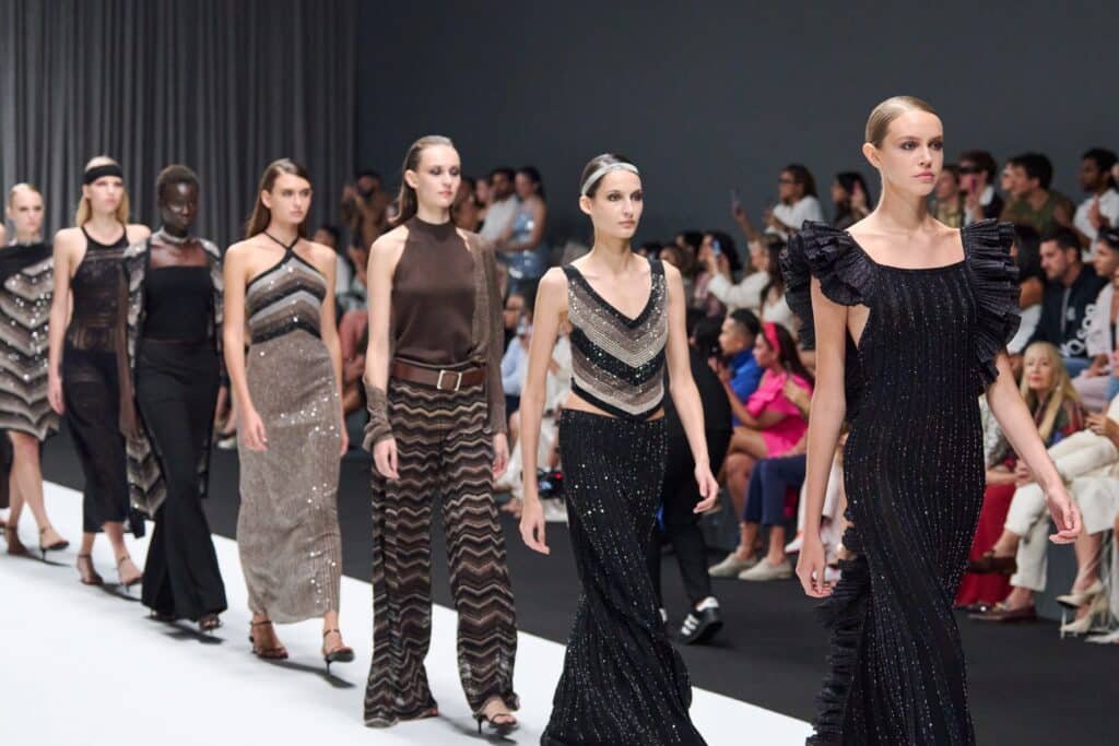 Models walk down a runway wearing elegant, sequined dresses in various dark shades.