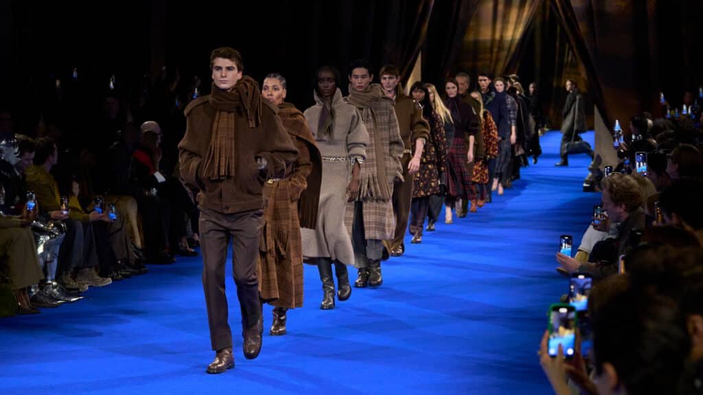 Models in brown plaid outfits walk down a blue runway during a fashion show, with an audience on either side.