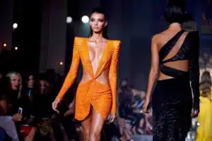 A model in an orange, textured dress with structured shoulders walks on a fashion runway beside another in a black dress.