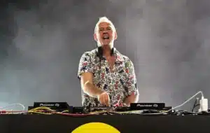 A DJ wearing a colorful shirt performs on stage, surrounded by sound equipment and lighting effects.