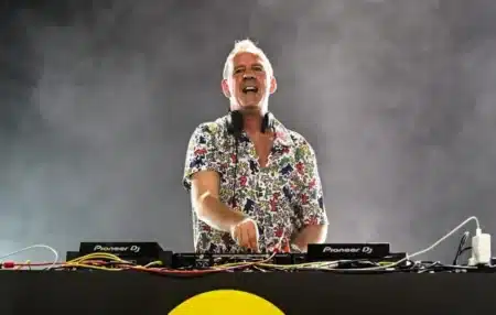 A DJ wearing a colorful shirt performs on stage, surrounded by sound equipment and lighting effects.
