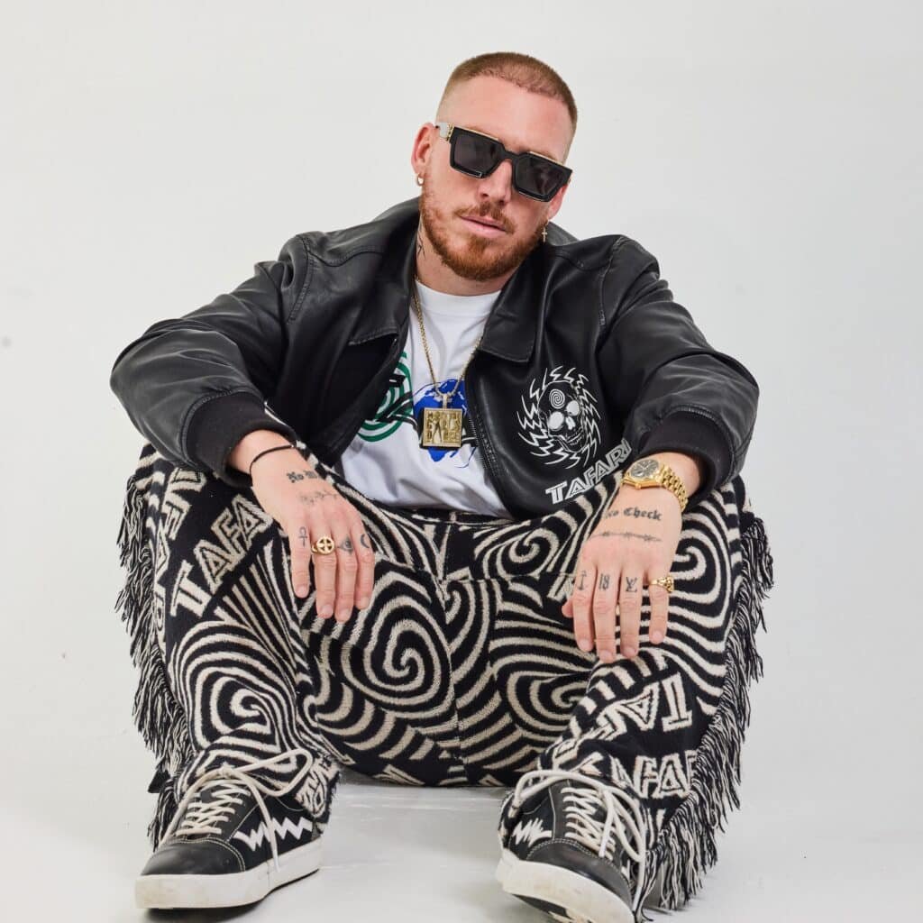 Person sitting on the floor wearing sunglasses, a black leather jacket, patterned pants, and black high-top sneakers.