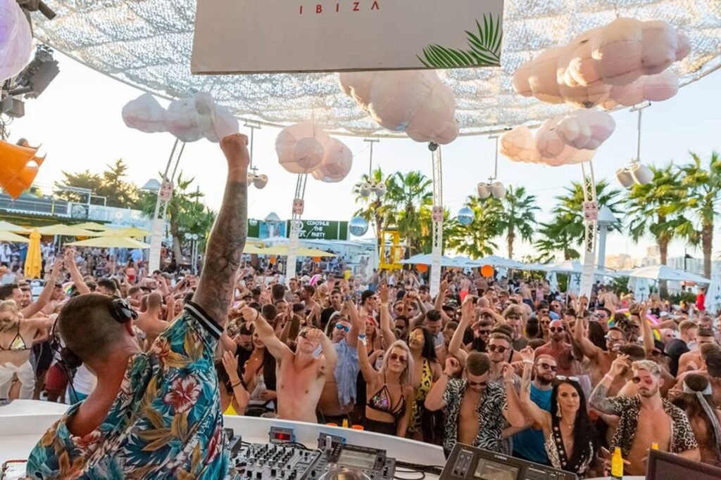 A DJ plays to a lively crowd at an outdoor party, with palm trees and decorative clouds overhead.
