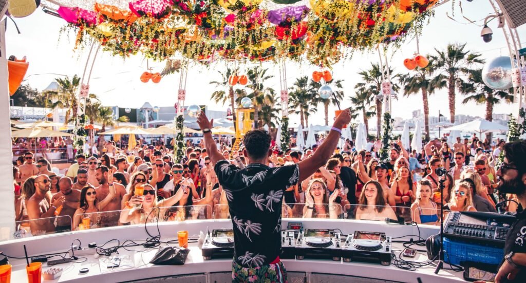 DJ playing at a vibrant outdoor beach party with a cheering crowd, tropical decor, and palm trees in the background.