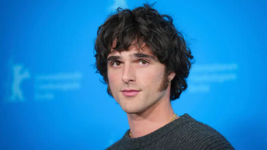 A person with curly dark hair and sideburns, wearing a chain necklace, stands against a blue backdrop with subtle text and logo.