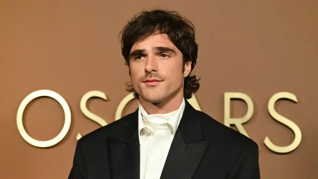 The Next James Bond with dark hair and a mustache wearing a white shirt and black jacket stands in front of a background featuring the word "OSCARS."