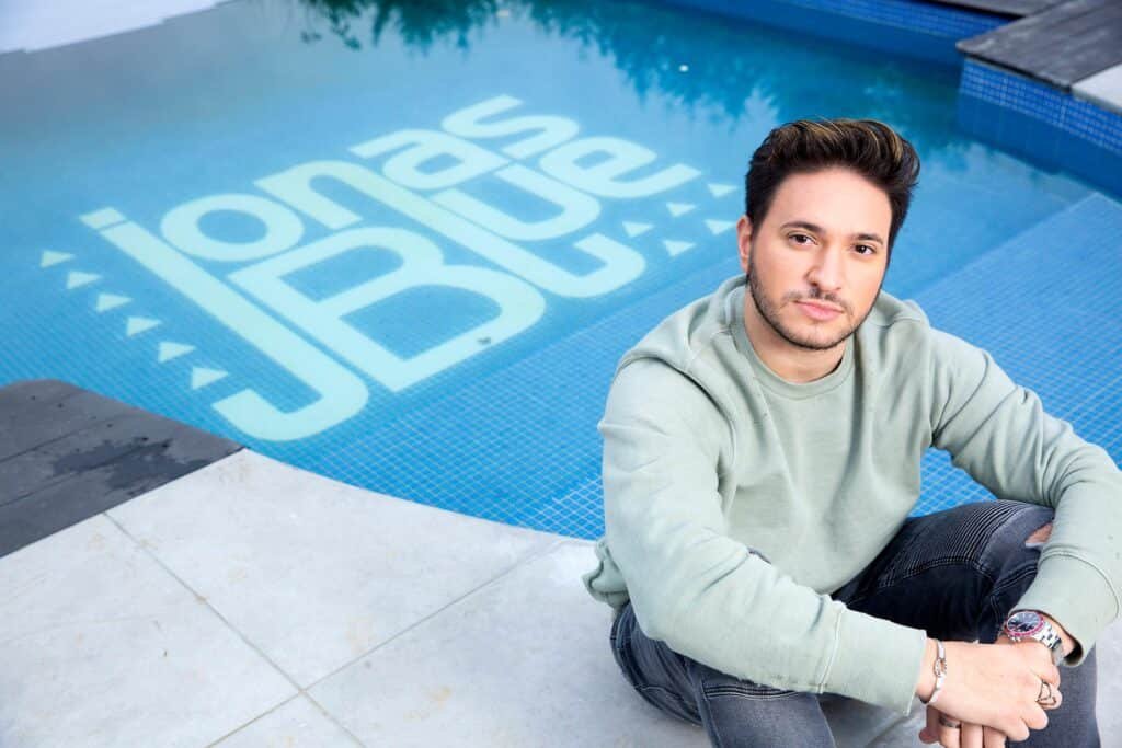 Jonas Blue in a green sweatshirt sits by a pool with "Jonas Blue" written on the pool floor.