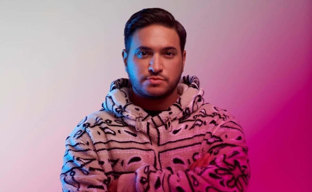 A person with short dark hair wearing a patterned hoodie stands against a gradient pink and purple background.