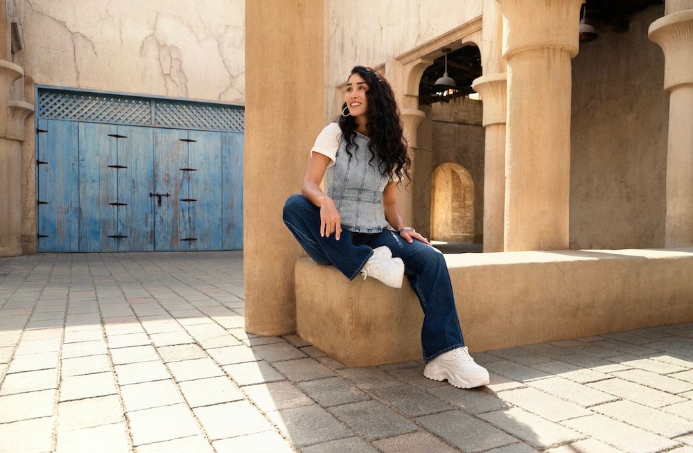 A person with long, dark hair wearing a gray vest and jeans sits on a low wall in a sunlit courtyard with stone arches and a blue door.