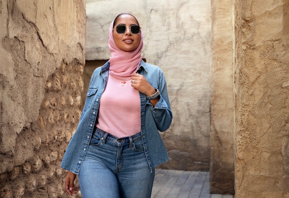 A woman wearing sunglasses, a pink hijab, a denim jacket, and jeans walks confidently down a narrow, rustic alleyway.