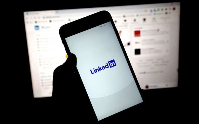 A person holding a smartphone displaying the LinkedIn logo, with a computer screen showing a LinkedIn webpage in the background.