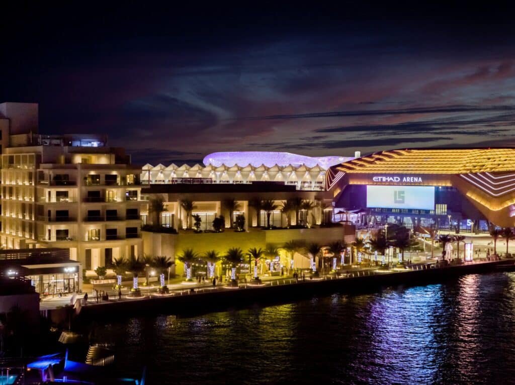 A night view of a waterfront with well-lit modern buildings, including the Etihad Arena, under a colorful sky.