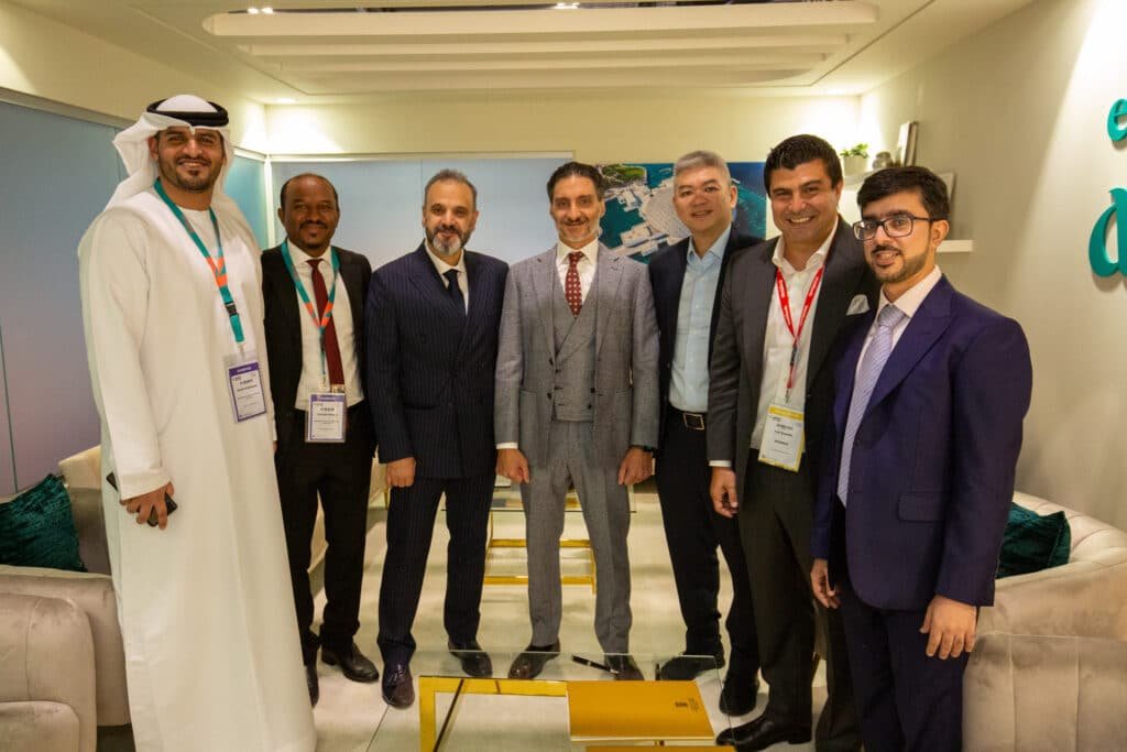 A group of seven men in business attire smiling at the Live Summit Middle East camera, standing in a modern, well-lit office setting.