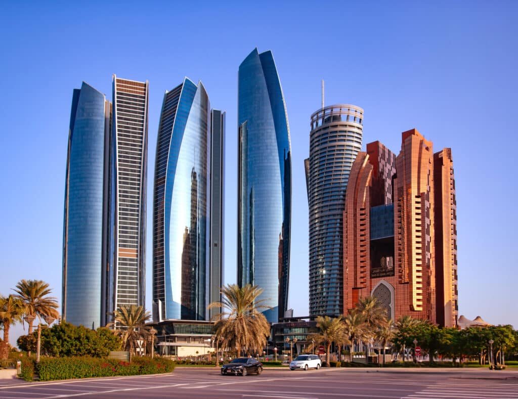 A group of modern skyscrapers with reflective glass facades set against a clear blue sky, surrounded by palm trees and a road.