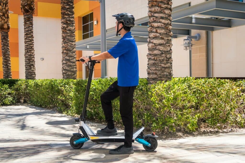 A person wearing a helmet stands on an electric scooter in a sunny outdoor area with palm trees and a building in the background.