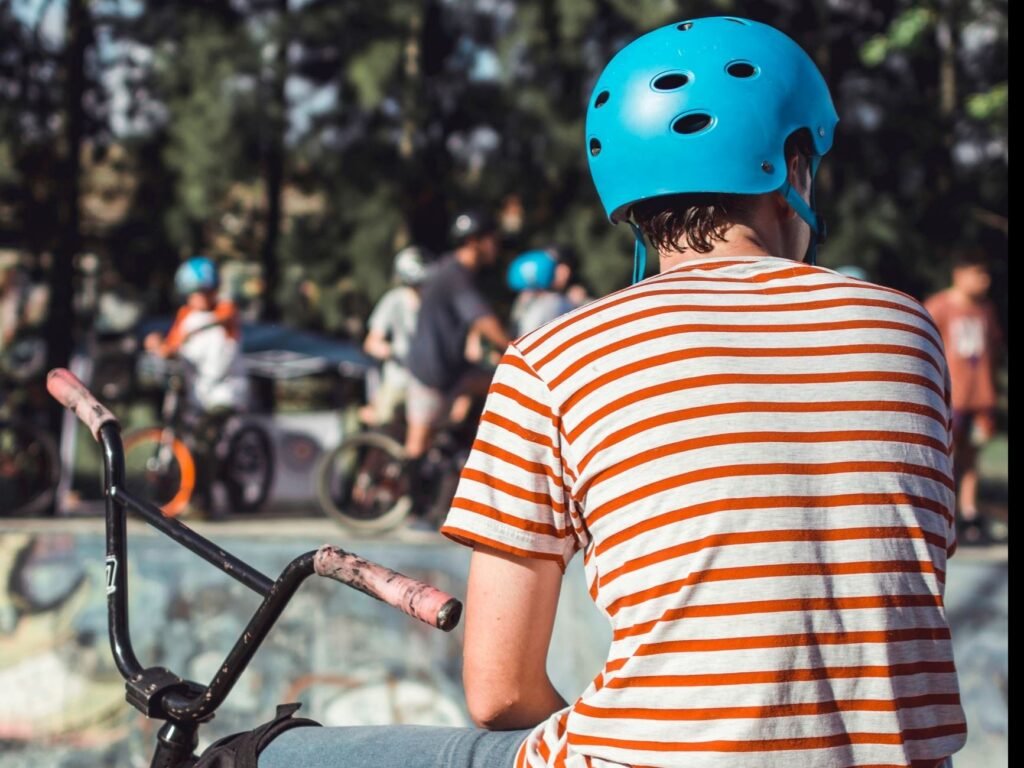 A person wearing a blue helmet and a striped shirt sits on a bicycle at a skate park with other people in the background.
