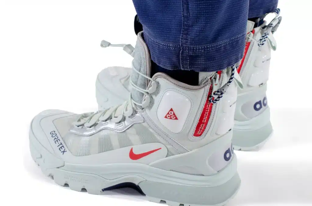 A person wearing white and gray Nike ACG Gore-Tex boots with red and blue accents and navy pants.