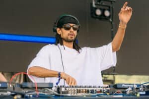 A DJ wearing sunglasses and a headband performs at a mixing console outdoors, with one hand raised.