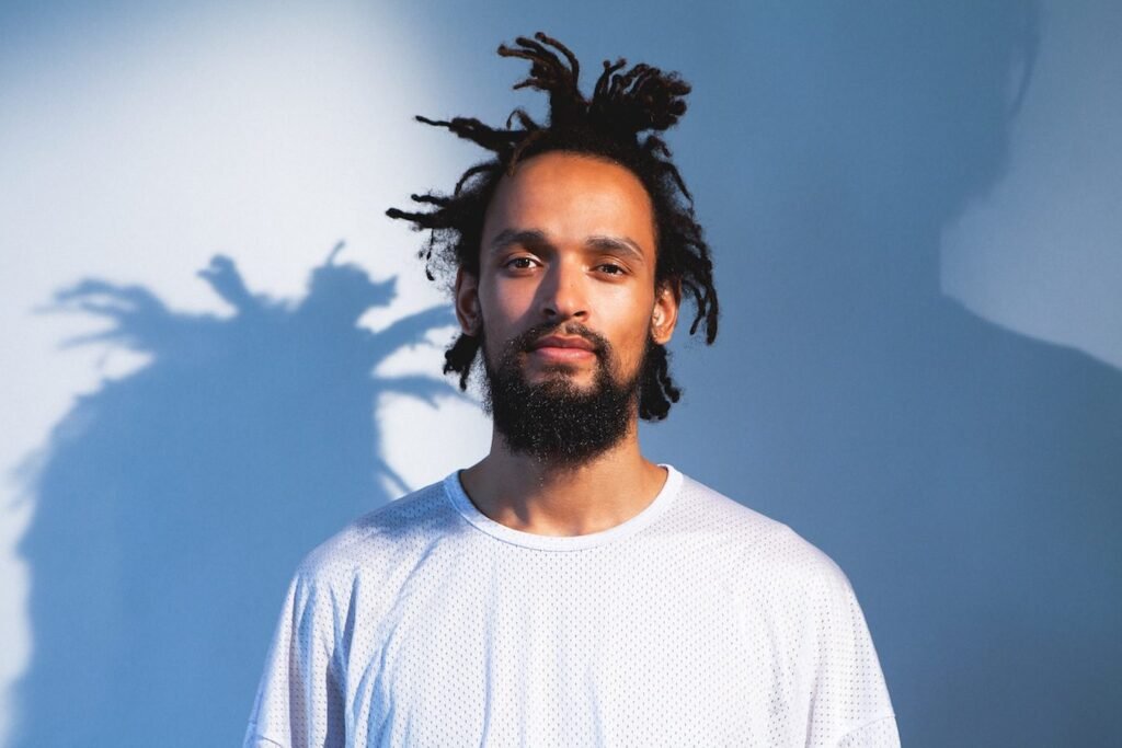 PAWSA Live at Be Beach with a beard and dreadlocks wearing a white shirt stands against a blue-lit background.