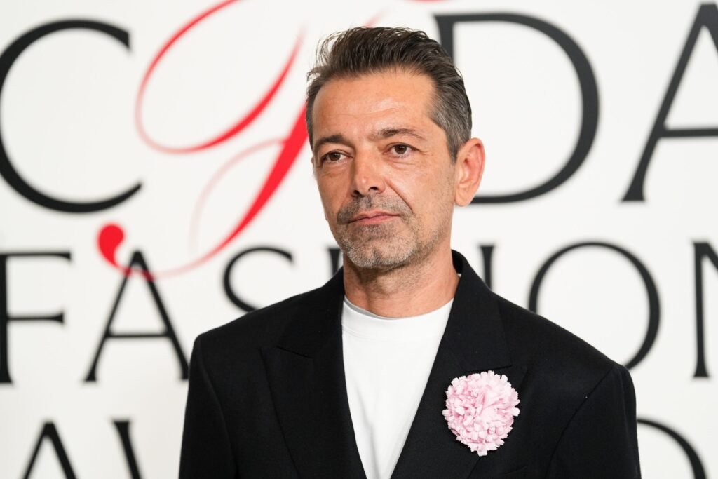 Pieter Mulier in a black suit with a pink flower pin stands against a background with large partial letters.