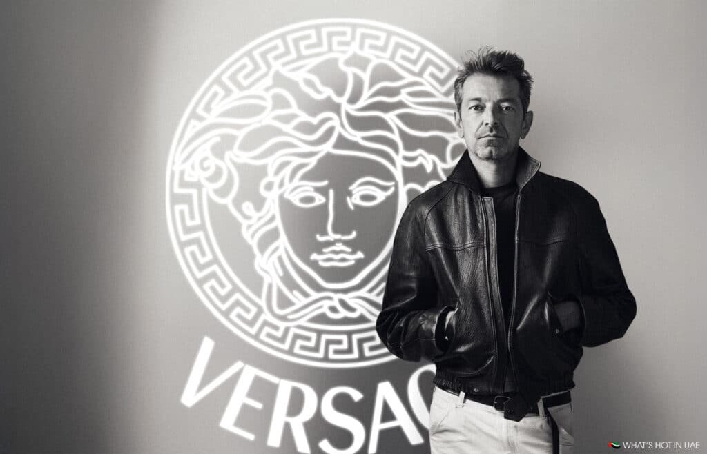 A person in a leather jacket stands in front of a Versace logo.