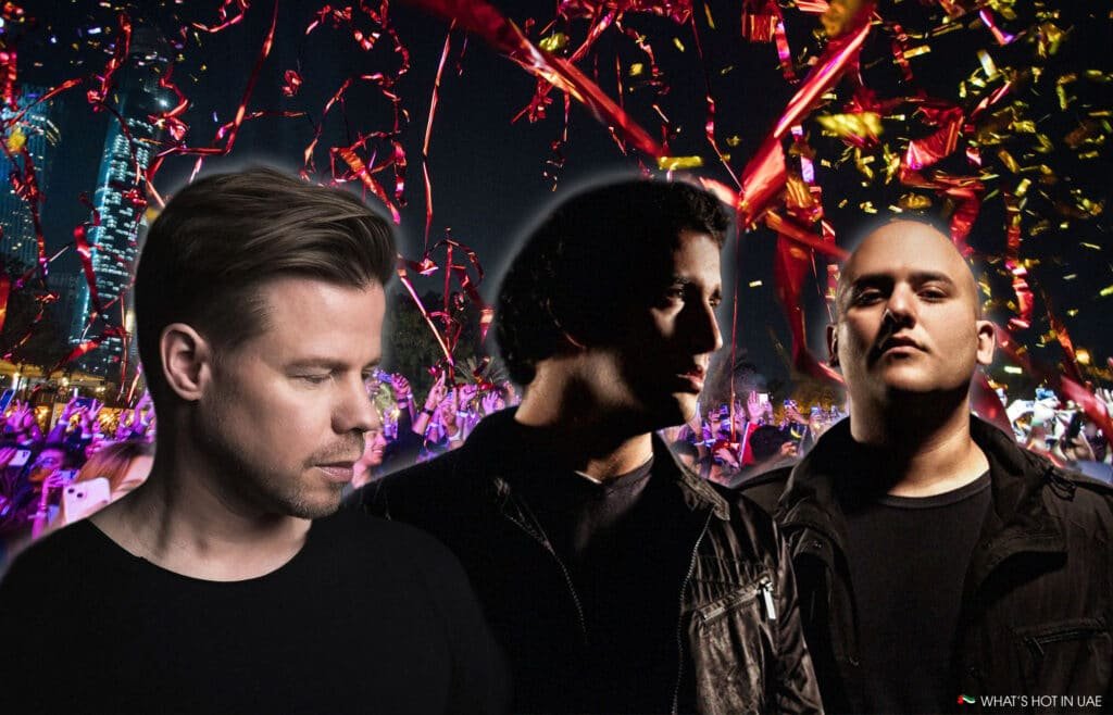 Three men in black clothing stand in front of a vibrant, colorful concert crowd with red and yellow streamers.