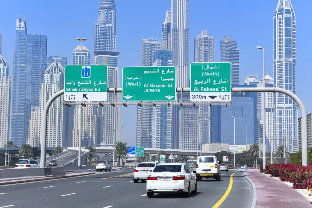 Ramadan Rush Hour Traffic Cars driving on a highway in a city with skyscrapers in the background and green road signs in Arabic and English overhead.
