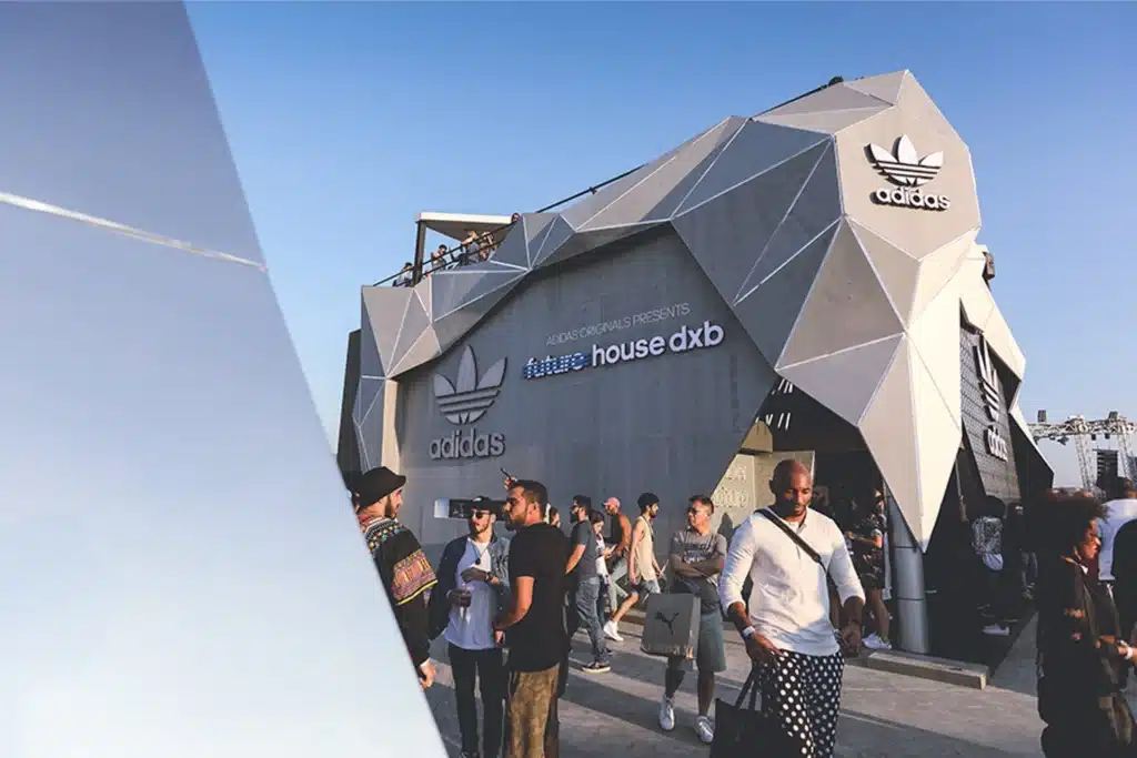 A modern, geometric Adidas building with people gathered outside, some carrying shopping bags, under a clear blue sky. Sneaker Culture in the UAE