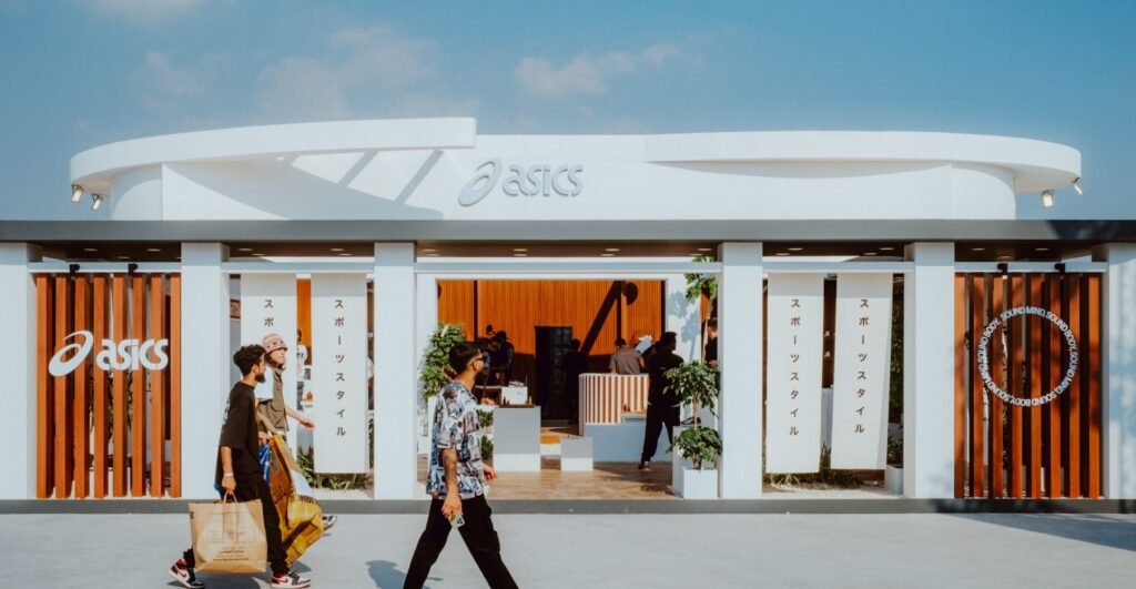 Two people walk past a modern, open-air ASICS store with wooden accents and Japanese text.