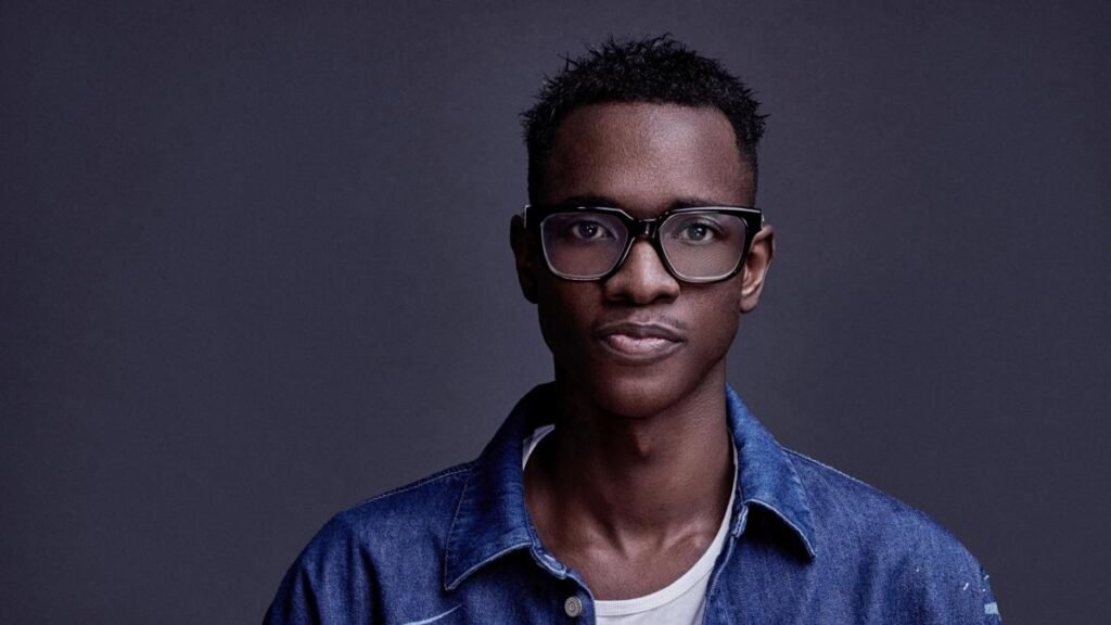SONA wearing glasses and a denim shirt stands against a dark background, looking directly at the camera.