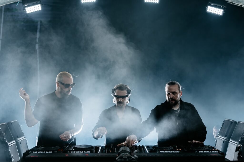 Swedish House Mafia wearing dark clothing perform with DJ equipment on a smoky stage.