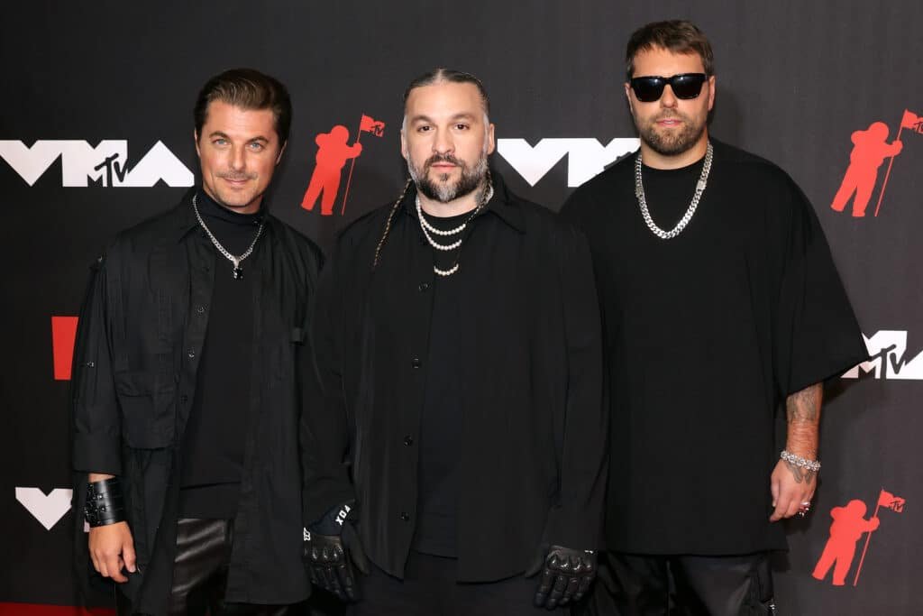 Swedish House Mafia dressed in black outfits pose on a red carpet with MTV VMA motifs in the background.