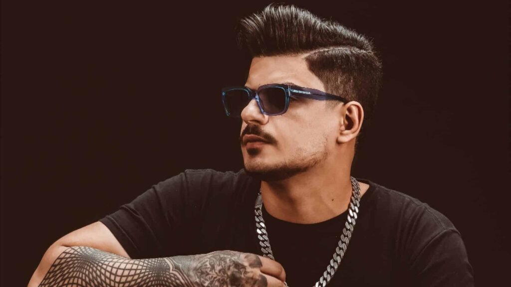 Vintage Culture with sunglasses, a stylish haircut, and tattoos on his arm wears a chain necklace against a dark background.