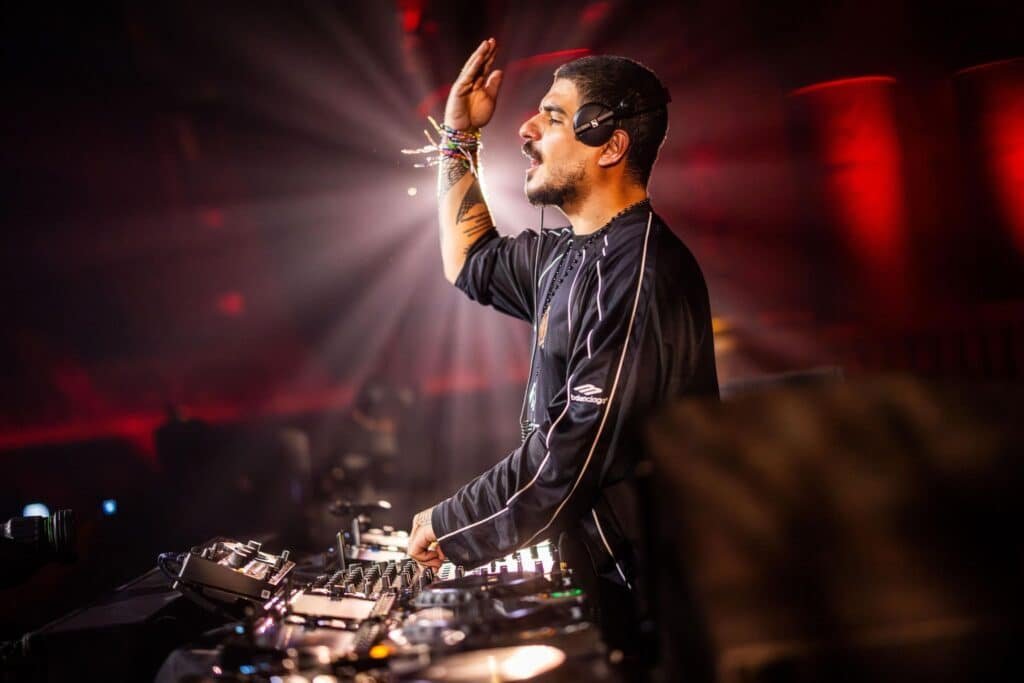 A DJ wearing headphones performs energetically with one hand raised, surrounded by sound equipment and colorful lights.